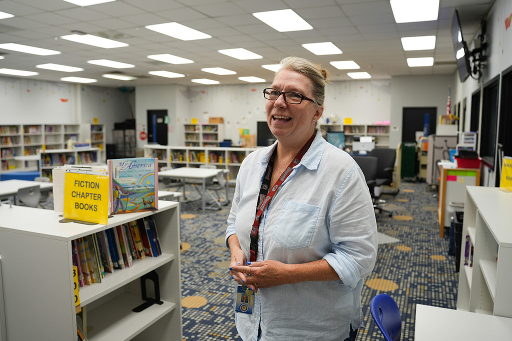 A woman wearing glasses and a blue shirt stands in a library with bookshelves and tables.