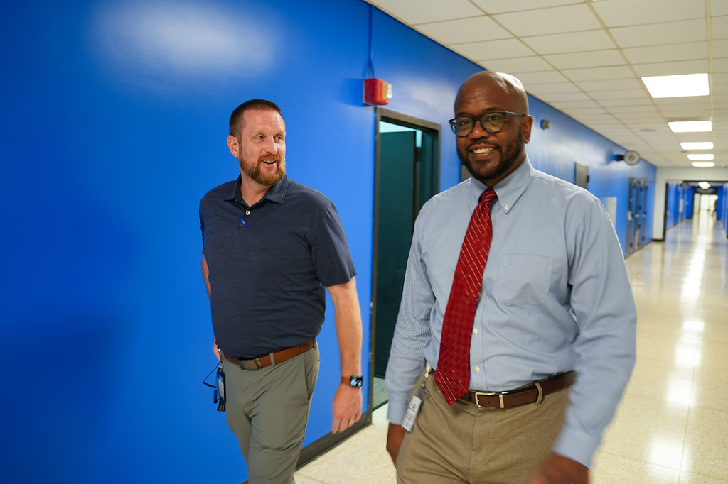 Two men in collared shirts and ties walk down a blue-walled corridor with doors on the left.