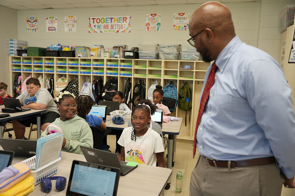A classroom with students working on laptops, a man in a blue shirt and red tie talks to a girl.