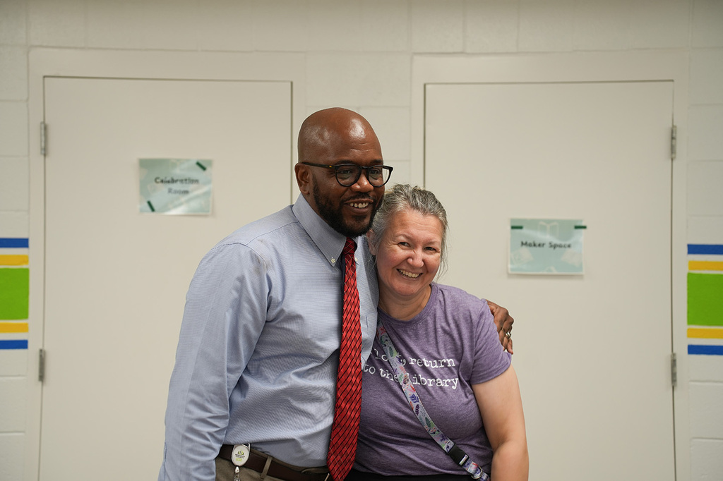 Man and woman standing and smiling, man in glasses and red tie, woman in purple shirt, white doors behind.