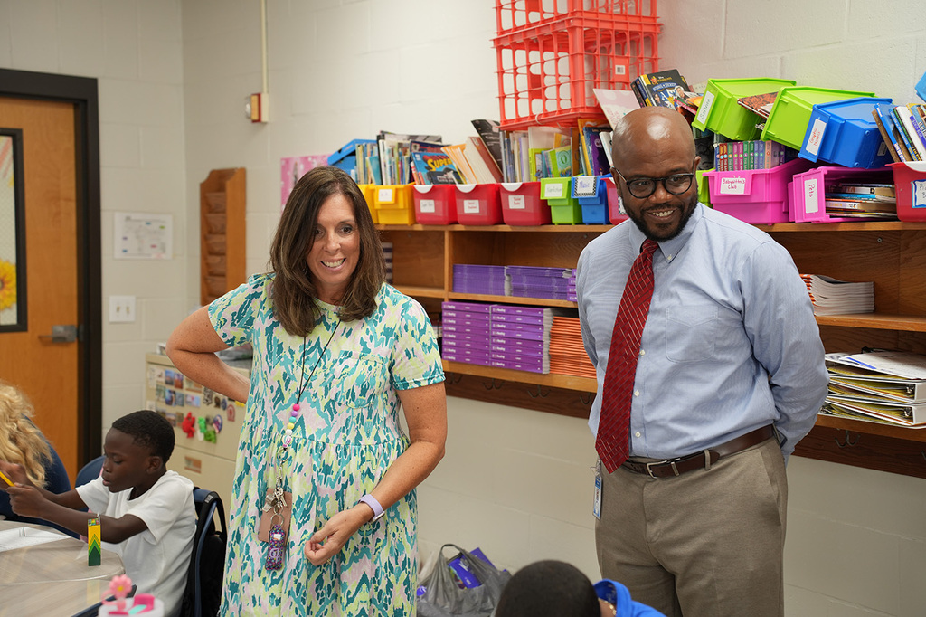 Two adults stand next to a shelf filled with school supplies. Behind them, children sit at tables.