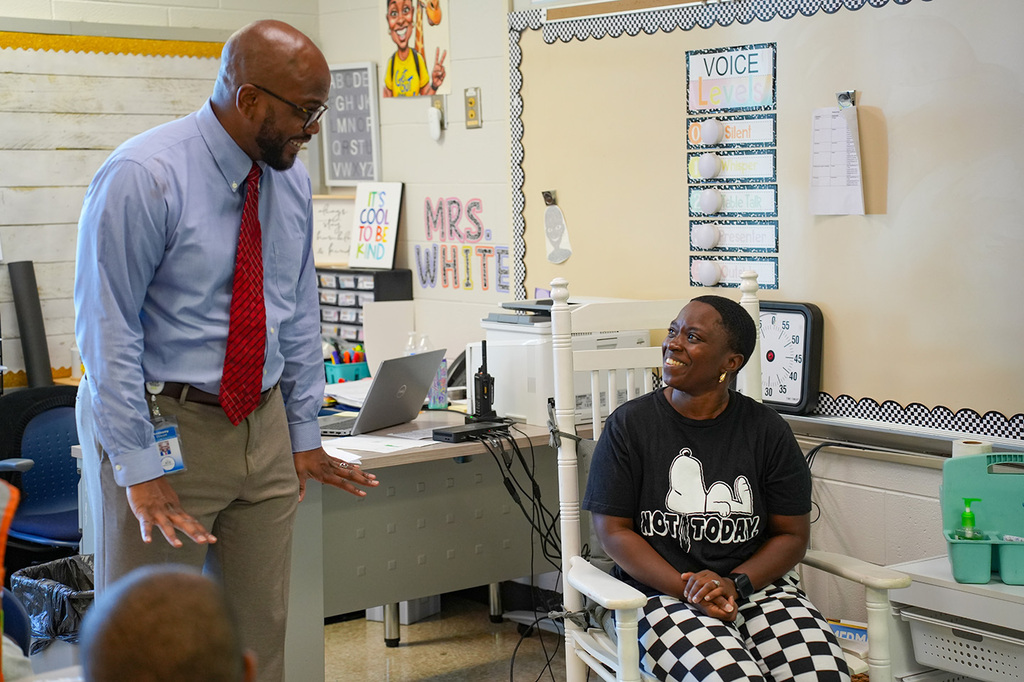 Man in blue shirt and red tie speaks to seated woman in black t-shirt in classroom.