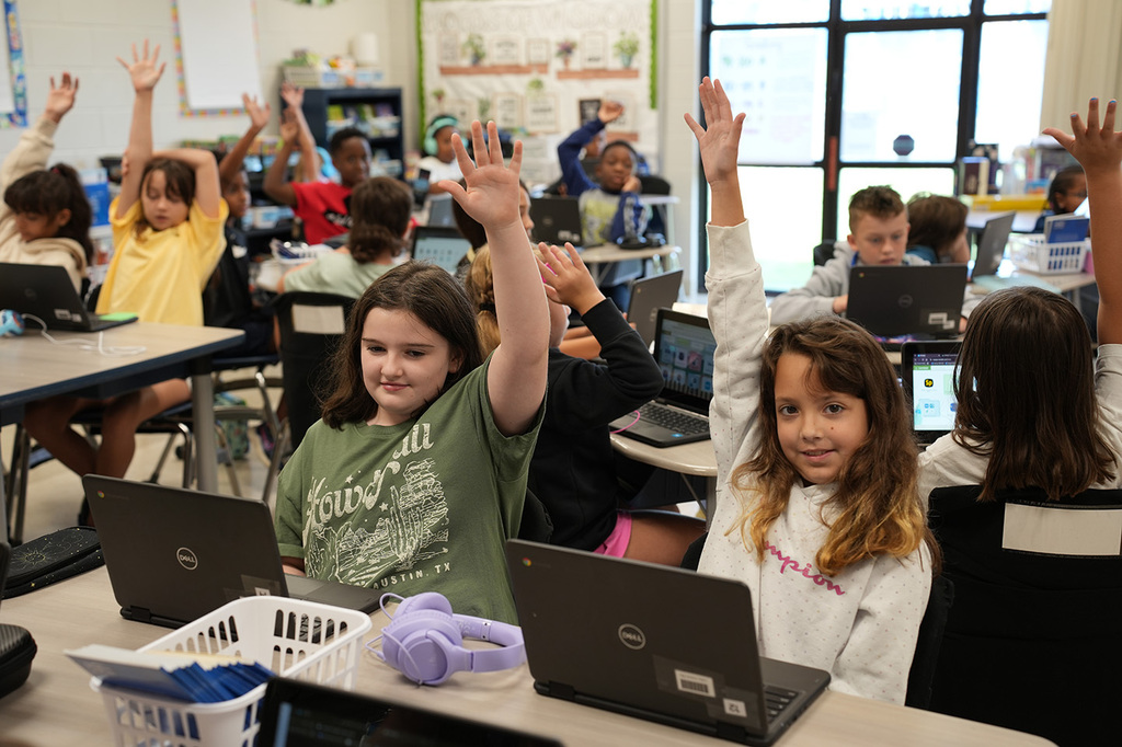 A classroom with children seated, raising hands; laptops on desks. Some wear yellow shirts, others long sleeves. Background shows windows and shelves with items.