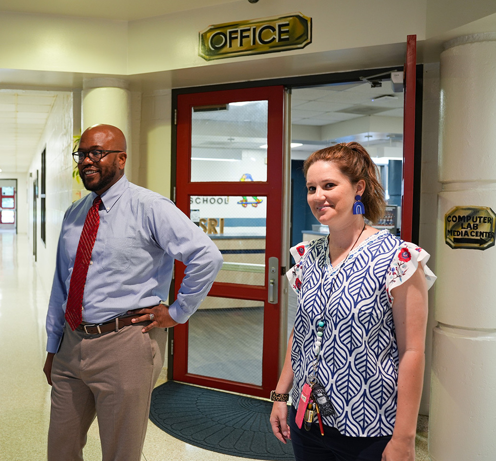 A man and a woman stand in front of an office door. The man has a red tie, the woman has earrings and a necklace.