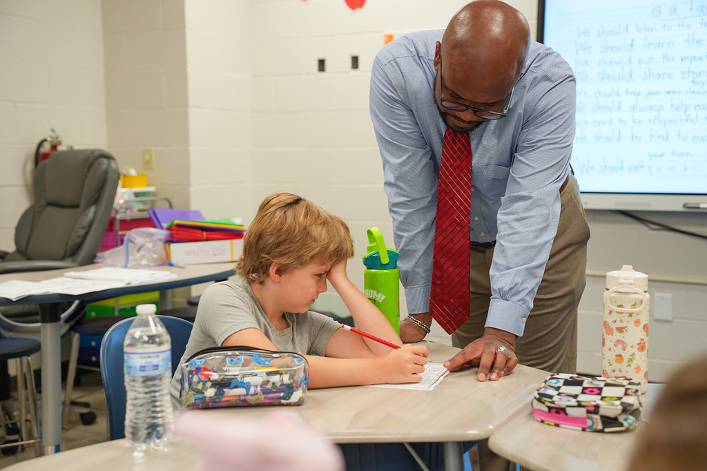 A teacher in glasses and a tie helps a student in a gray shirt with a writing assignment.