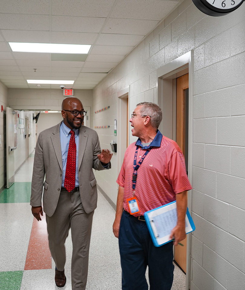Two men in suits, one with glasses, walk in a hallway. One holds a folder and wears a lanyard.