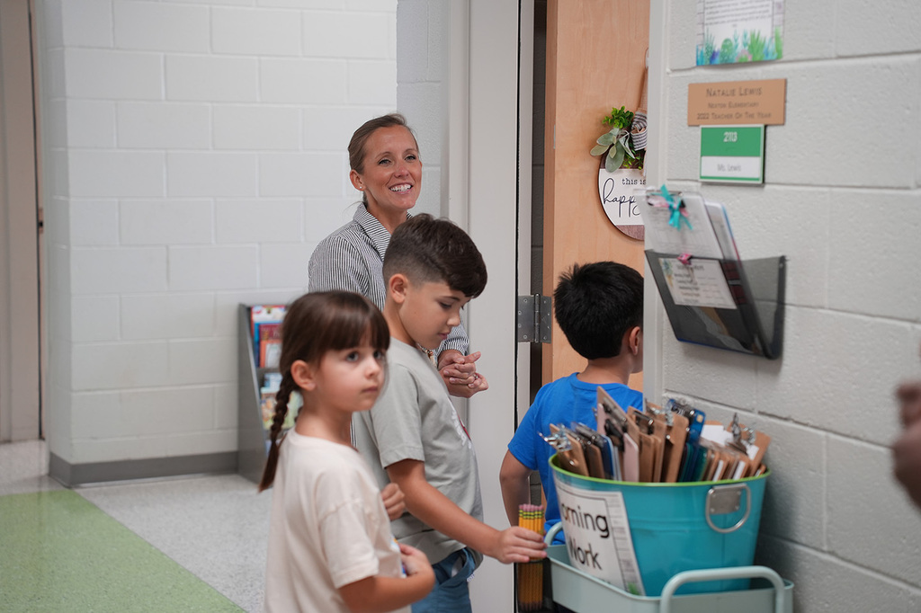 Three children and an adult look at a blue container with colorful straws and papers. A wall with bulletin boards is behind them.