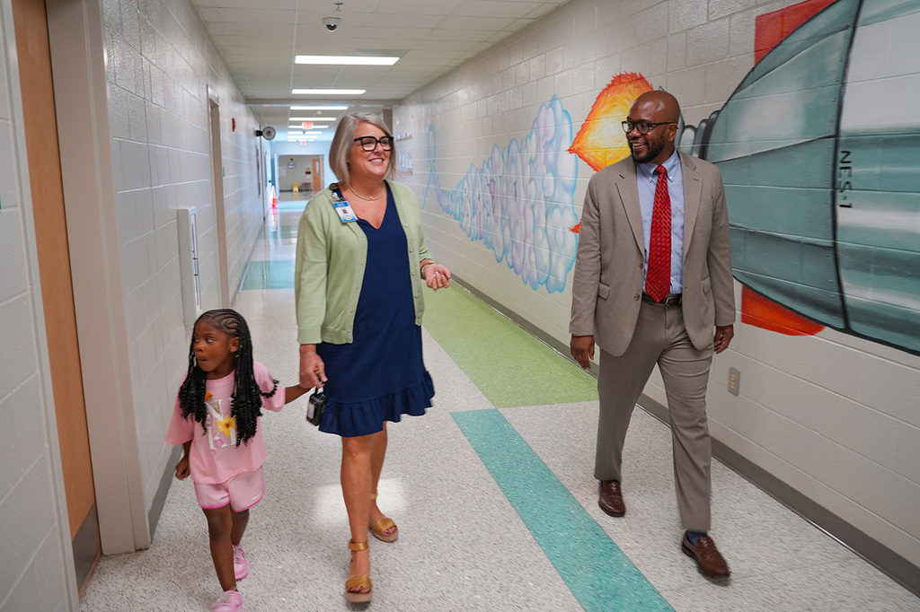 A woman and a man, holding a young girl's hand, walk through a brightly lit hallway with colorful murals.