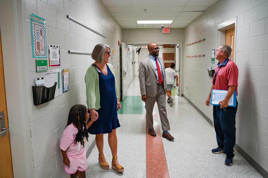 Four adults and a young girl in a school hallway, facing each other with a young child in the background.