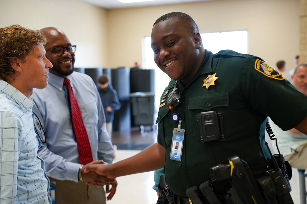 A uniformed officer shakes hands with a man in a light blue shirt and red tie in an indoor area.