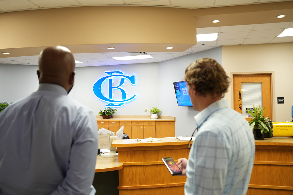 Two individuals face away from a camera in an office. A lighted emblem is on the wall.