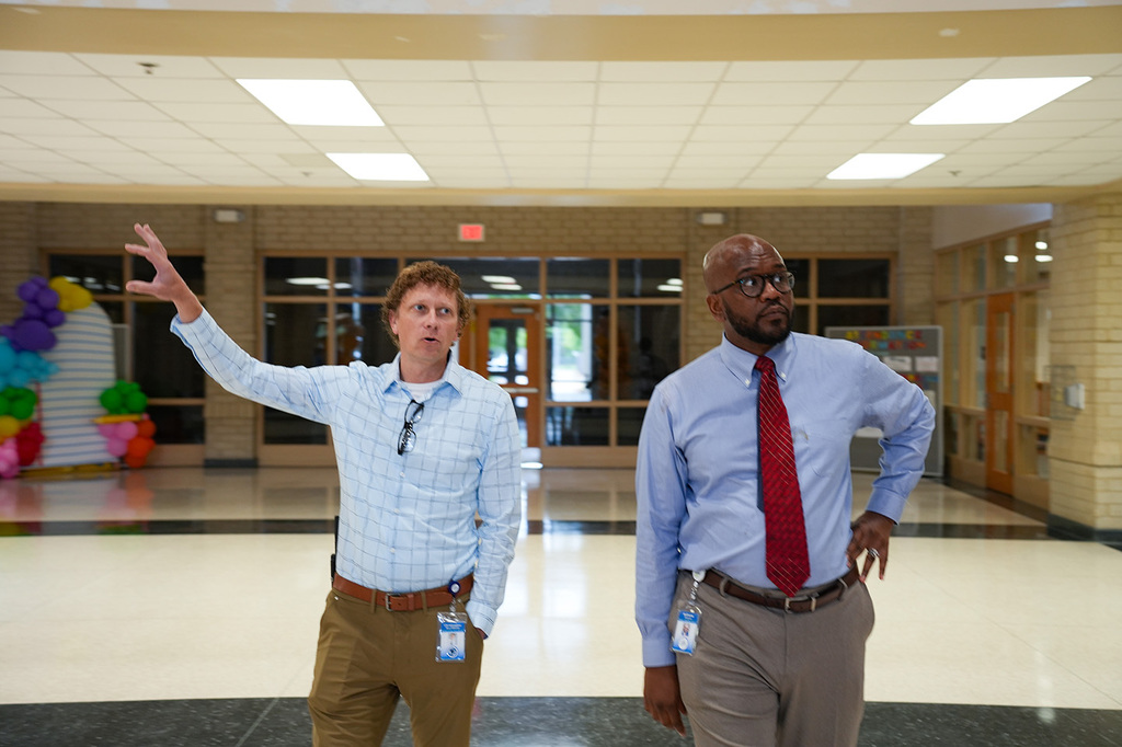 Two men in formal attire, one gesturing, stand in a school hallway with decorations and glass doors.