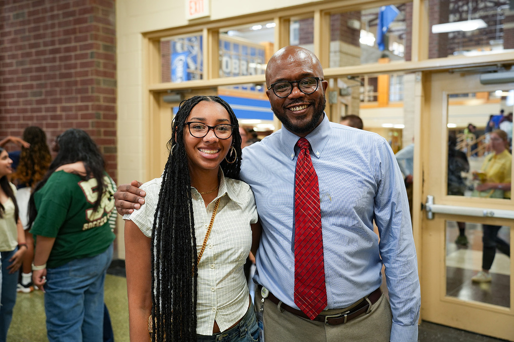 A man and a woman pose for a photo in a school hallway with brick walls and glass doors.