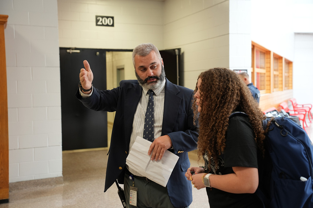 Man in a suit and tie speaks to a woman with curly hair and a backpack.