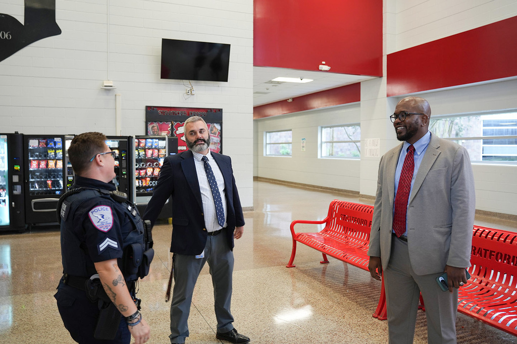 Three men stand in a hallway with red benches, vending machines, and a TV on the wall.