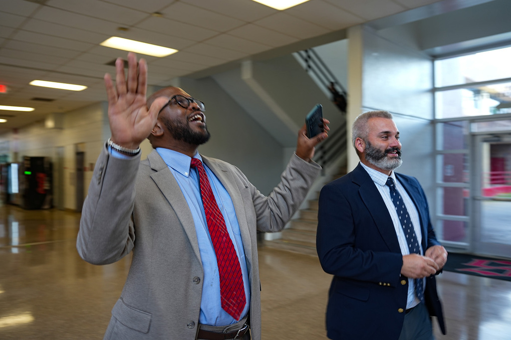 Two men in suits and ties inside a building with one raising his hand and the other holding a phone.