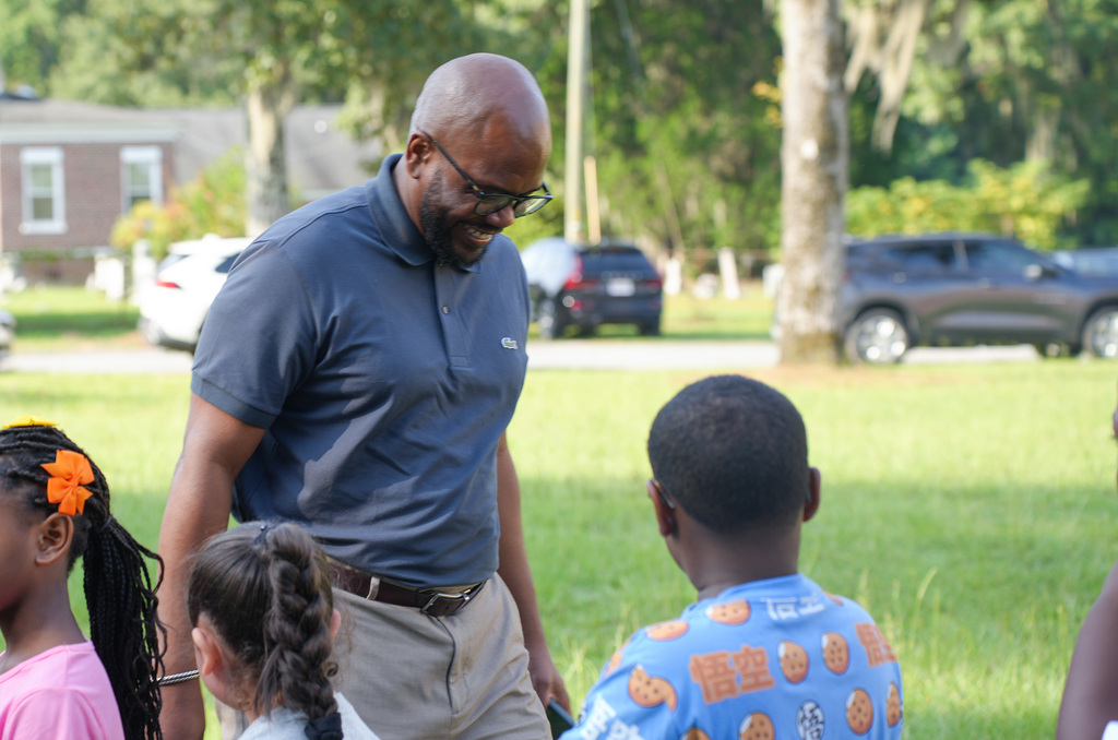 A man in glasses and a blue polo stands in a grassy area with children. Behind him, houses and cars are visible.