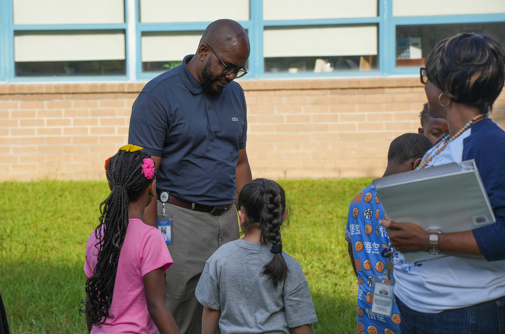 Adults and children stand outdoors, facing each other. A brick school building with windows is in the background.