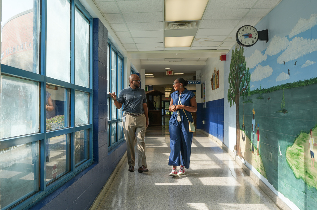Two people are standing in a hallway, discussing. One wears a blue jumpsuit and the other a gray shirt. A mural decorates the wall.