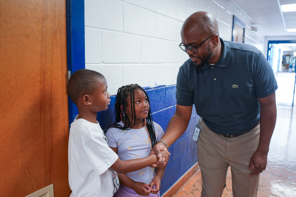 A man with glasses holds the hands of two children in a school hallway with blue and white walls.