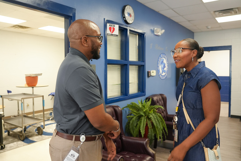 Two adults stand in a room. A man wears glasses and a badge. A woman wears glasses and a blue dress.