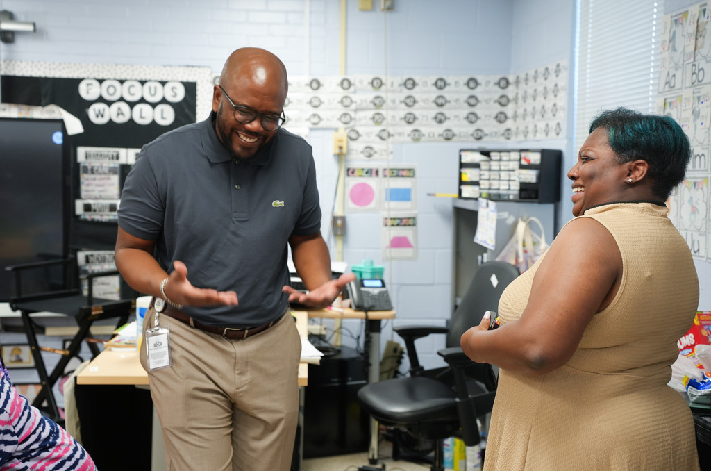A man in glasses and a woman in a sleeveless top converse in a classroom. A computer and chairs are behind them.