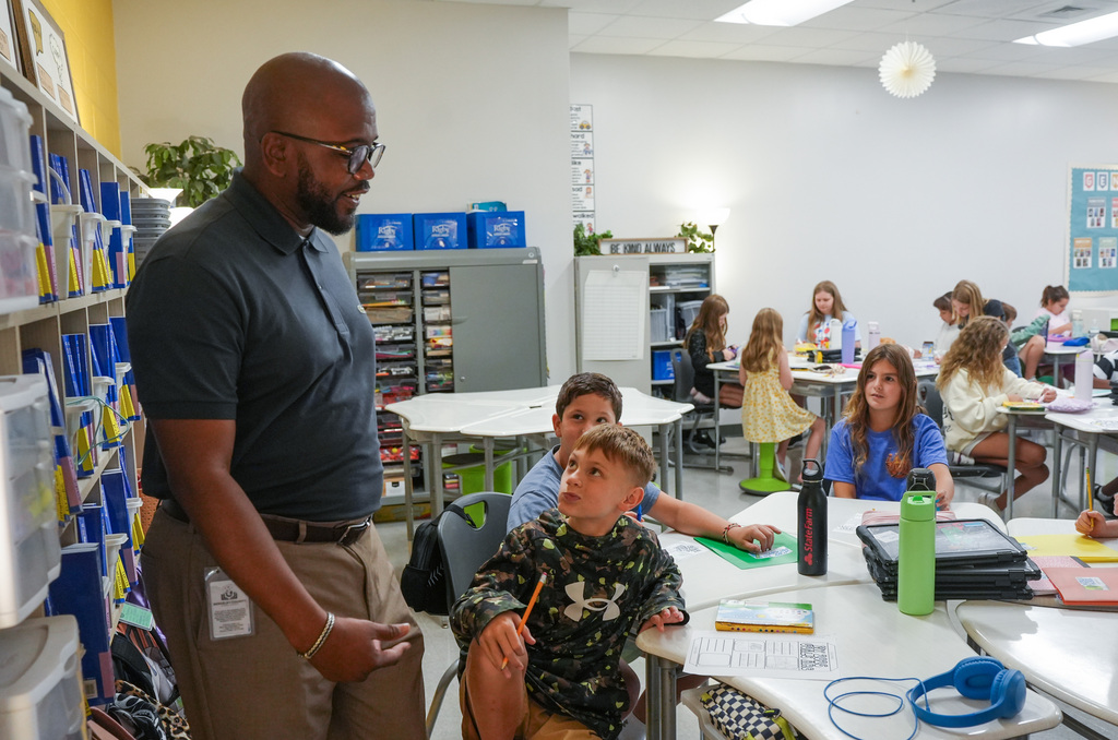 An adult male with glasses stands in a classroom, talking to a child seated at a desk.