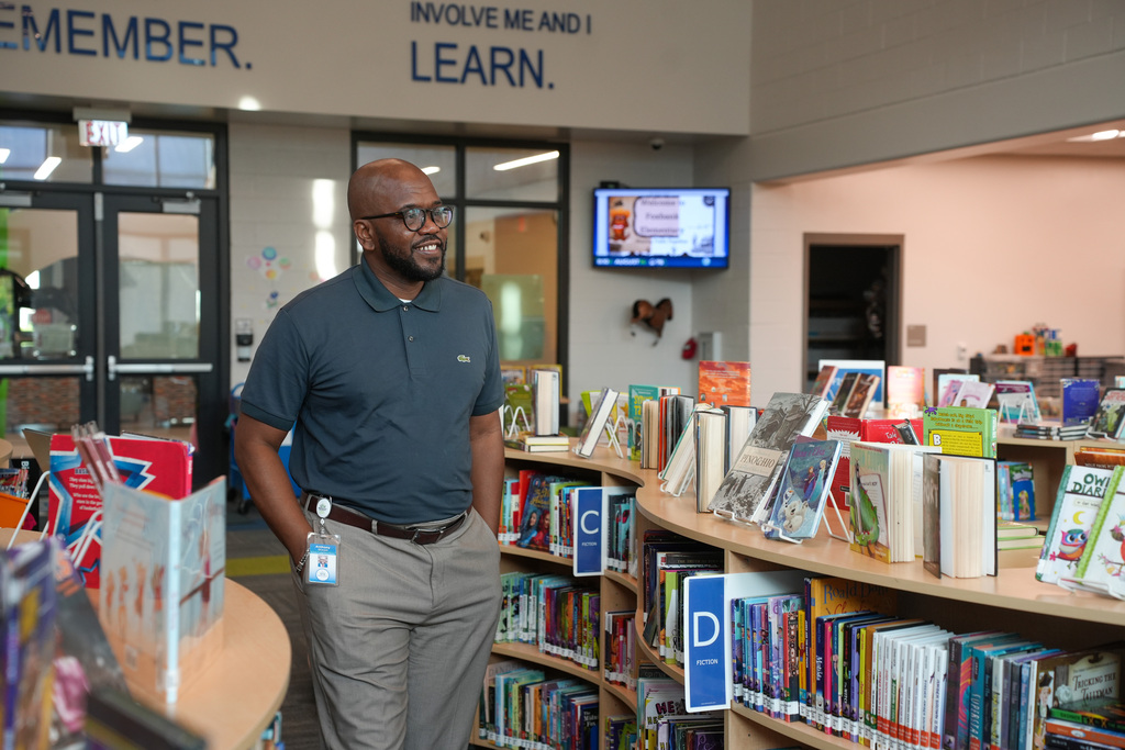 A man stands in a library with shelves filled with books, wearing a blue shirt and gray pants.