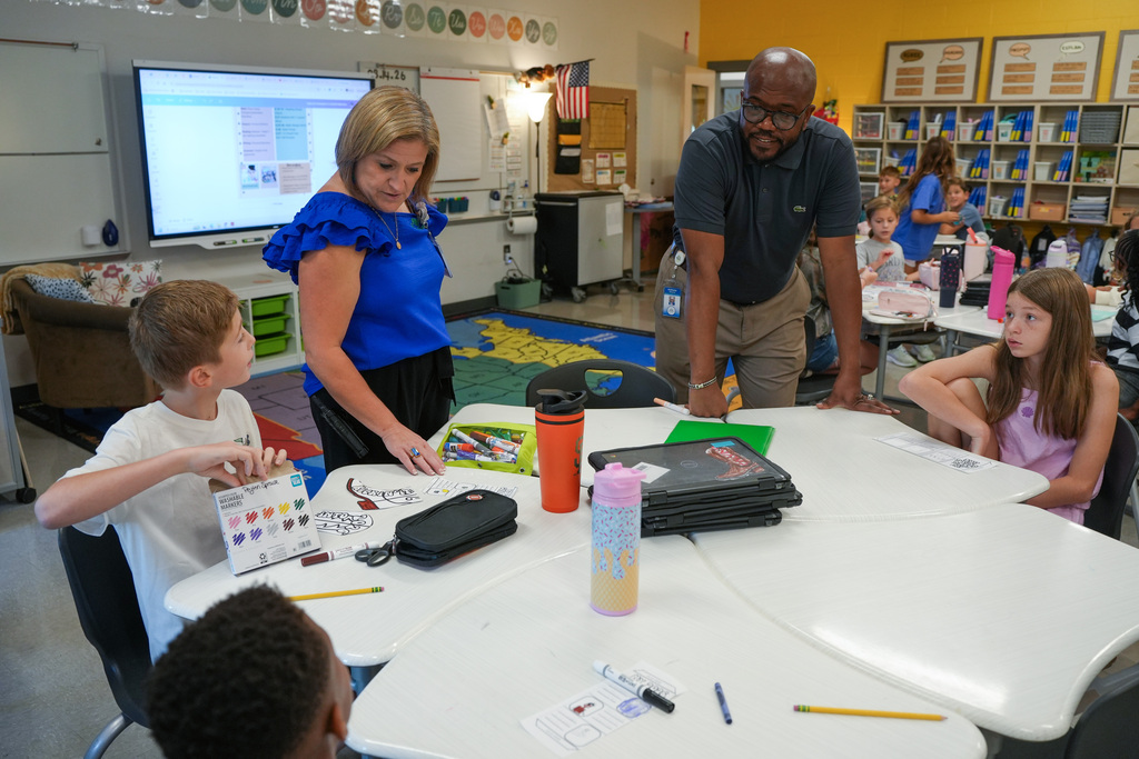 Several children are seated at a table. A woman and a man are standing behind them.