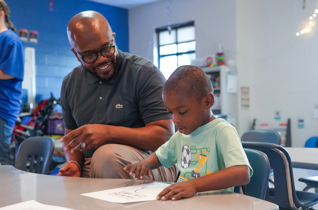 A man and a child sit at a table, the child drawing on paper. The man wears glasses and a dark shirt, the child a light green shirt.