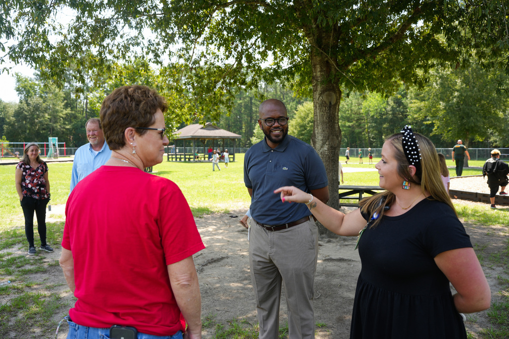 A group of people in a park. A man and a woman converse, while another woman in a red shirt listens.