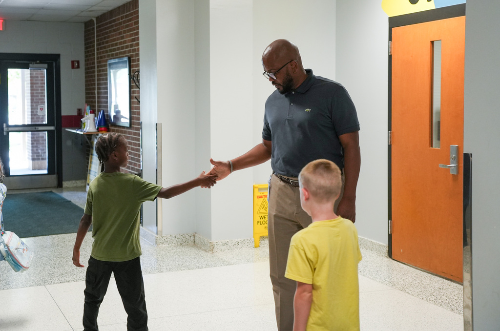 Two boys greet a man in a school hallway, one boy wearing green and the other yellow.