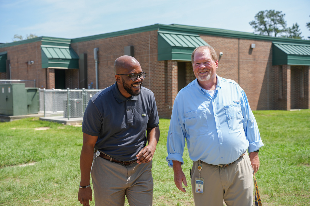 Two men stand outside a brick building with a green roof, one in a blue shirt and the other in gray.