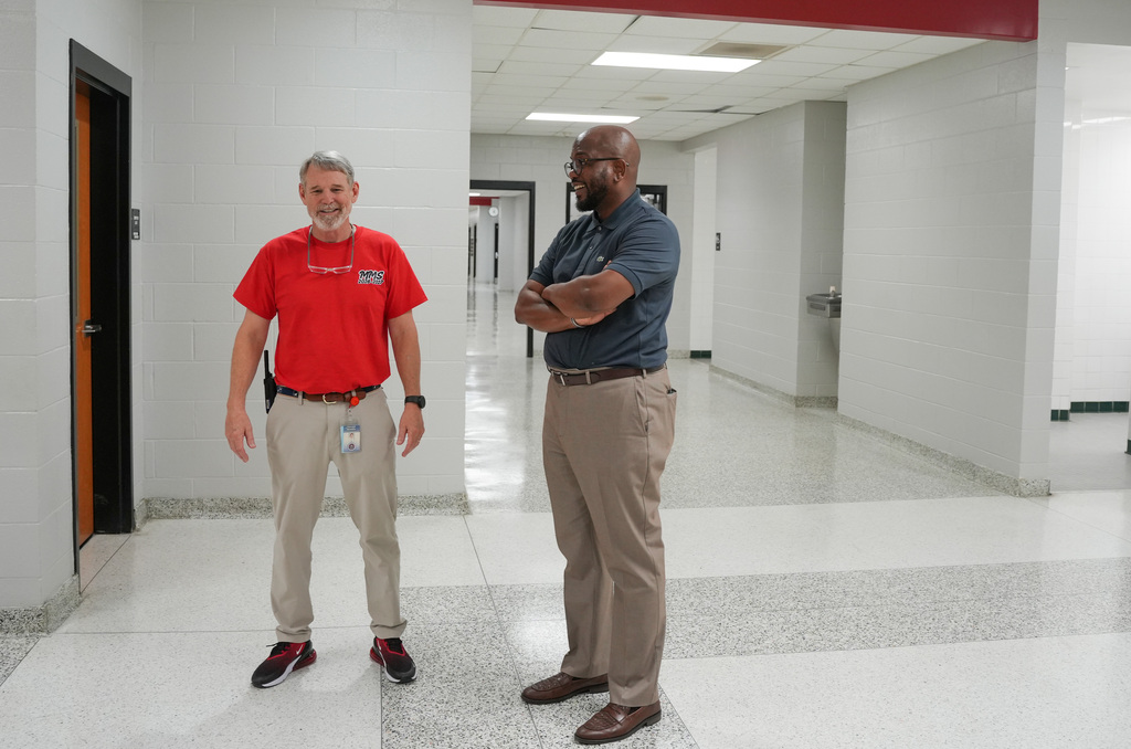 Two men, one in a red shirt and the other in a blue shirt, stand in a hallway with white walls and polished floors.