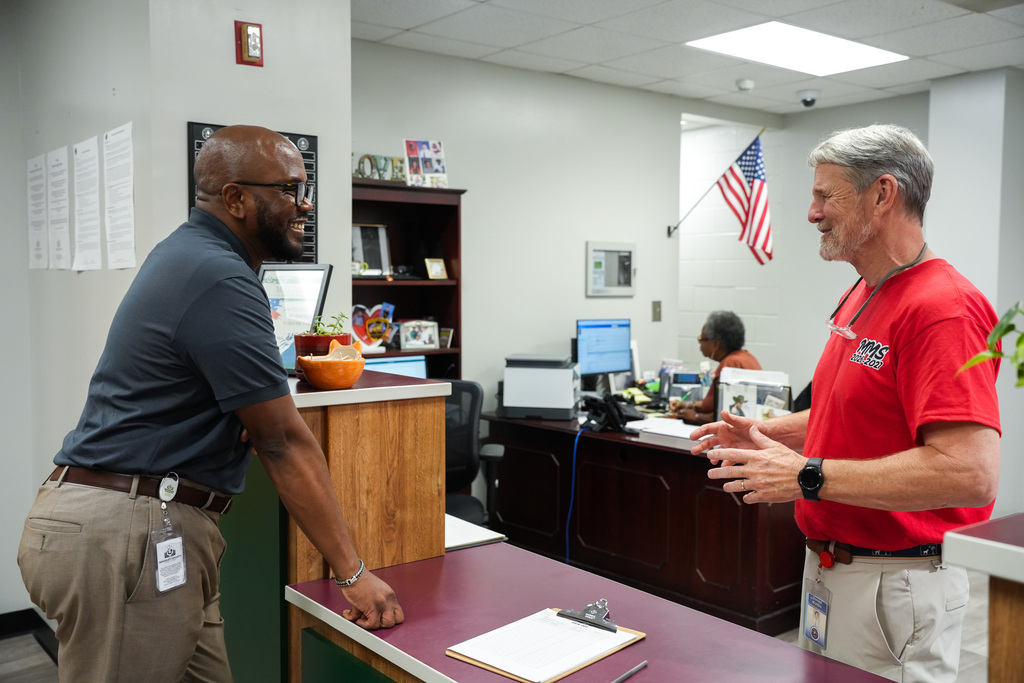 Two men conversing in an office with monitors, desks, and an American flag in the background.