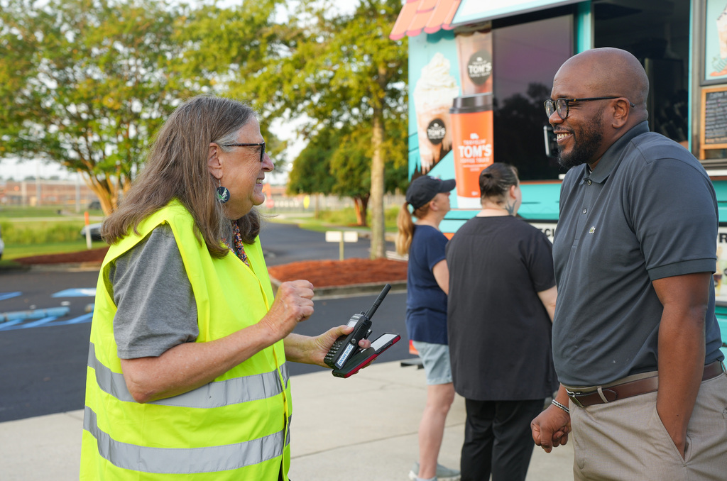 A woman in a yellow vest speaks with a man near a food truck; others stand nearby.