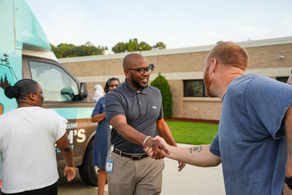 Four people shake hands in front of a vehicle and building. Two men shake hands.