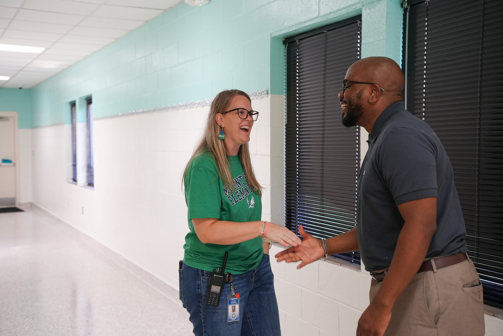 Two individuals converse in a hallway with white walls, blue accents, and windows with blinds.