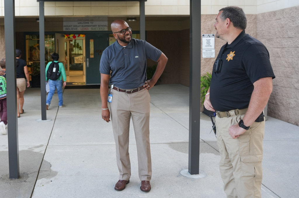 Two men converse outside a building with a stone wall, metal poles, and a wet floor.
