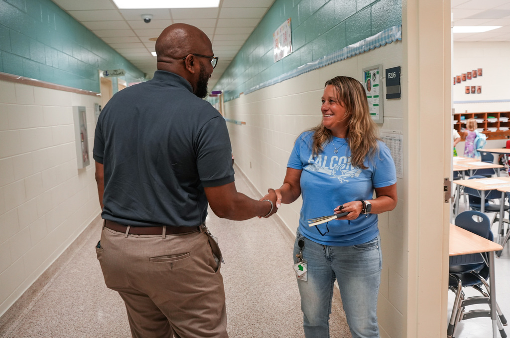 A man and a woman shake hands in a hallway, with the woman holding a key. They stand in front of a classroom.