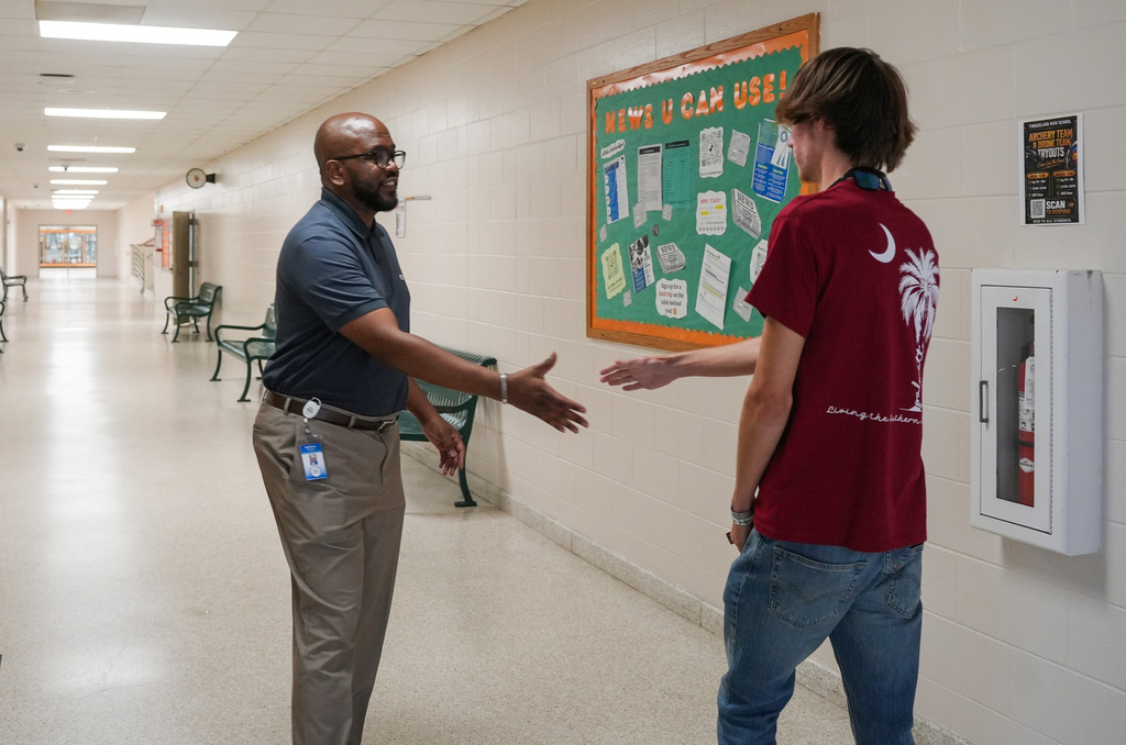 Two men face each other in a hallway. One wears glasses and a name tag, the other a maroon shirt. Behind them, a bulletin board and fire extinguisher.