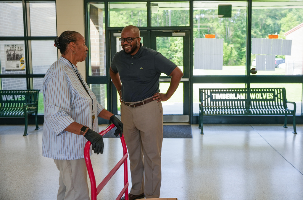 Two people in a hallway; one holds a red cart, and the other wears a belt. Benches are behind them.