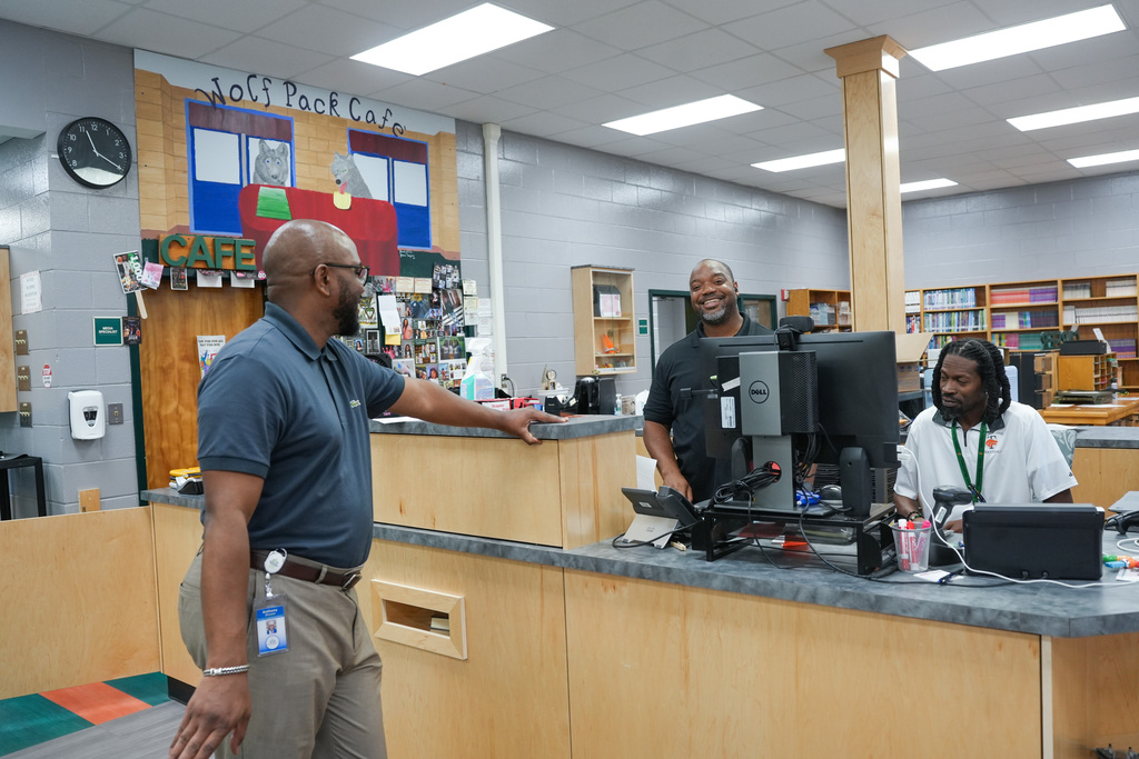 Three men in an office. A man in a light blue shirt stands behind a counter. Another man sits at a computer.