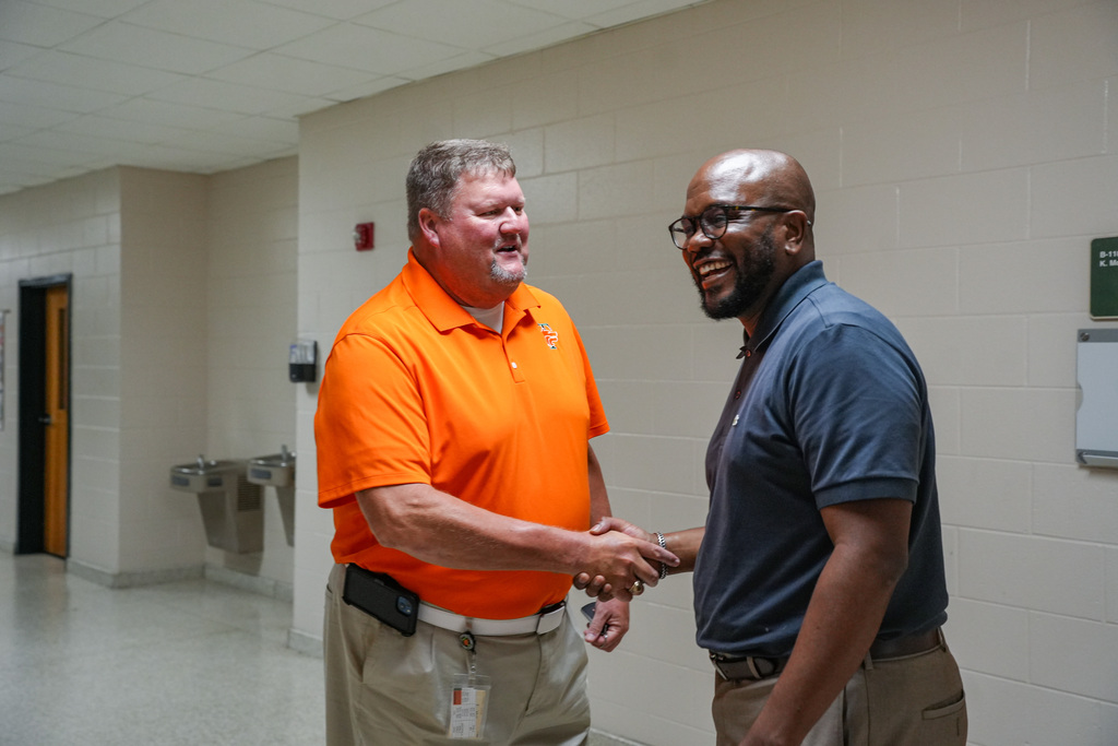 Two men in a hallway shake hands. One wears an orange shirt, the other a blue shirt. Behind them, a white wall has a door and a sink.