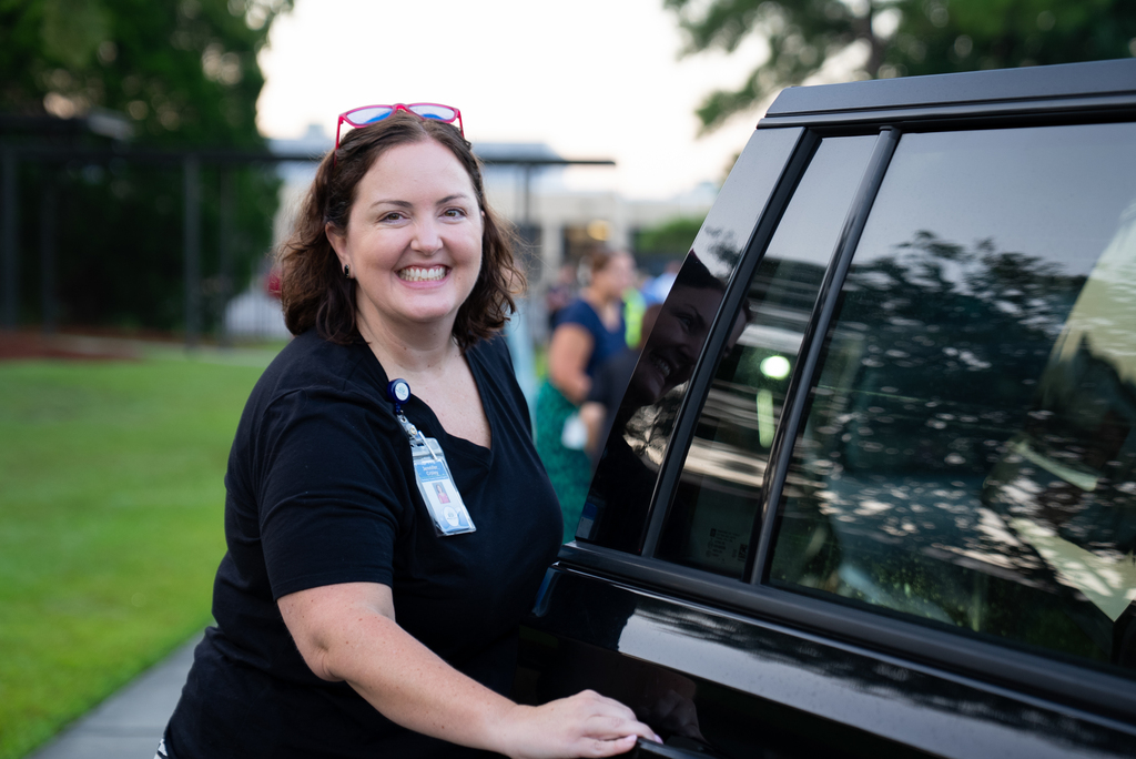 A smiling woman, wearing a black shirt and ID badge, stands next to a black vehicle on a sidewalk.