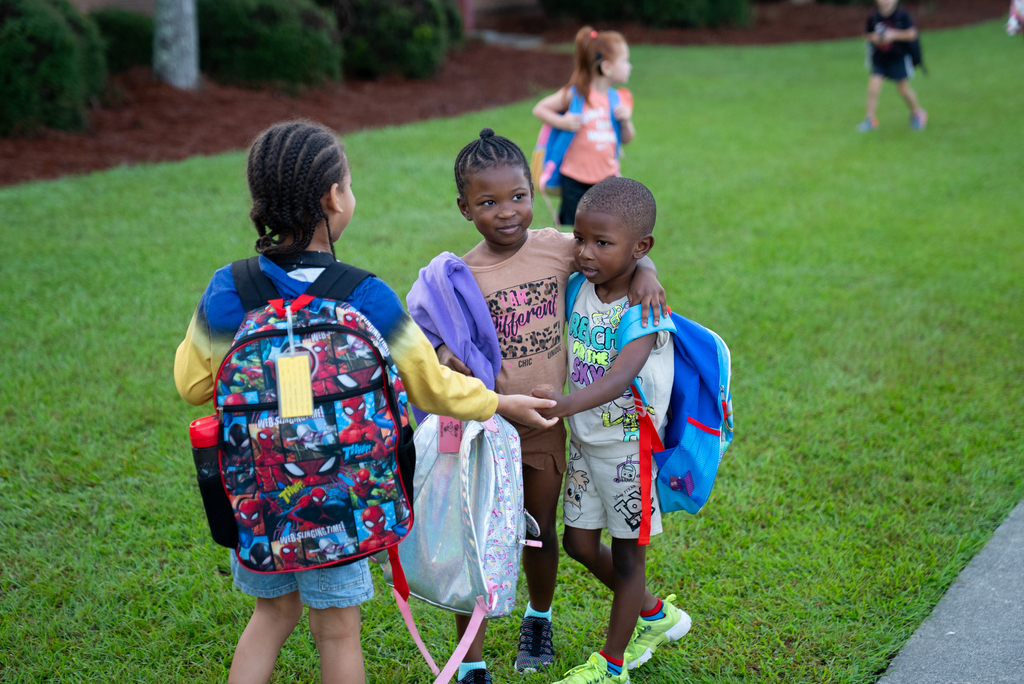 Three children stand together on grass with backpacks. One holds another's arm. A sidewalk and more children are in the background.