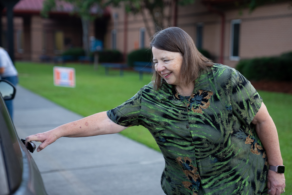 Woman with short hair and a camouflage shirt opening the door of a vehicle outside.