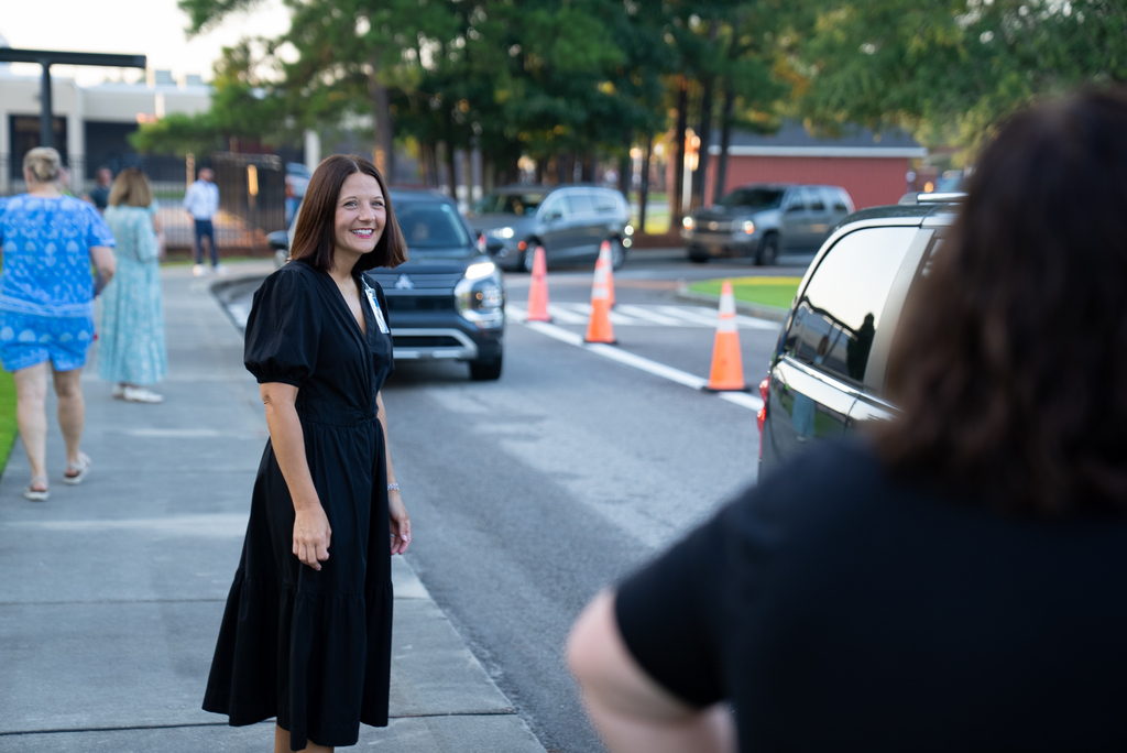A woman in a black dress stands on a sidewalk, facing a parked car and a person in a black shirt.