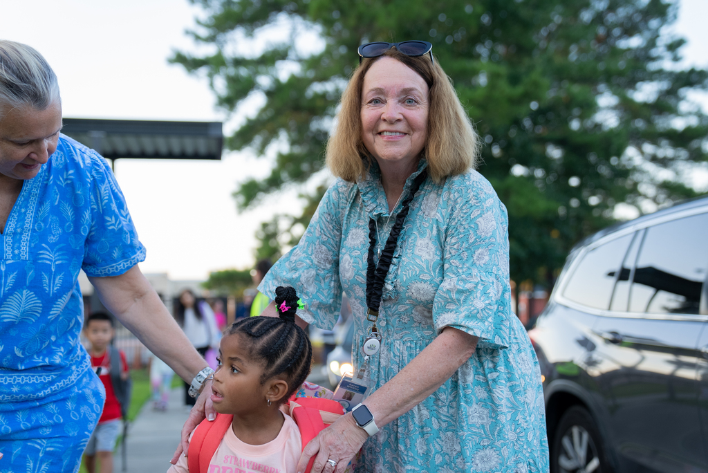 Woman in blue scrubs holds young girl in pink shirt, both smiling. A parked car is in the background.