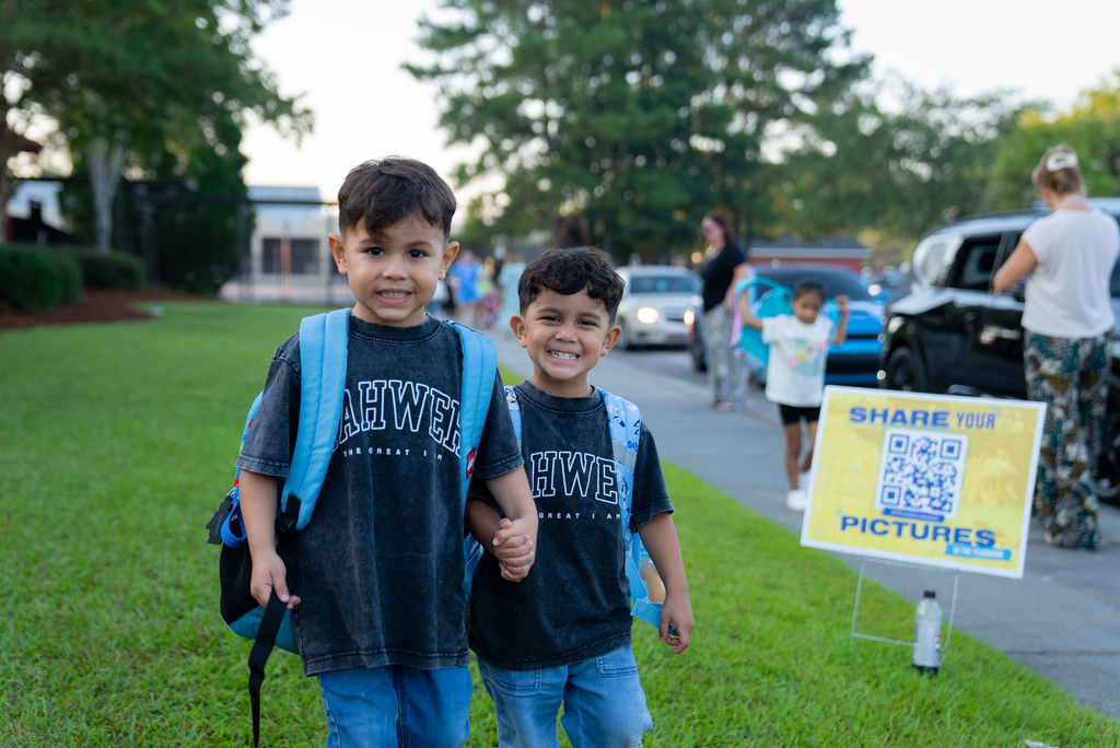 Two smiling young children in school uniforms and backpacks hold hands near a sign with a QR code.